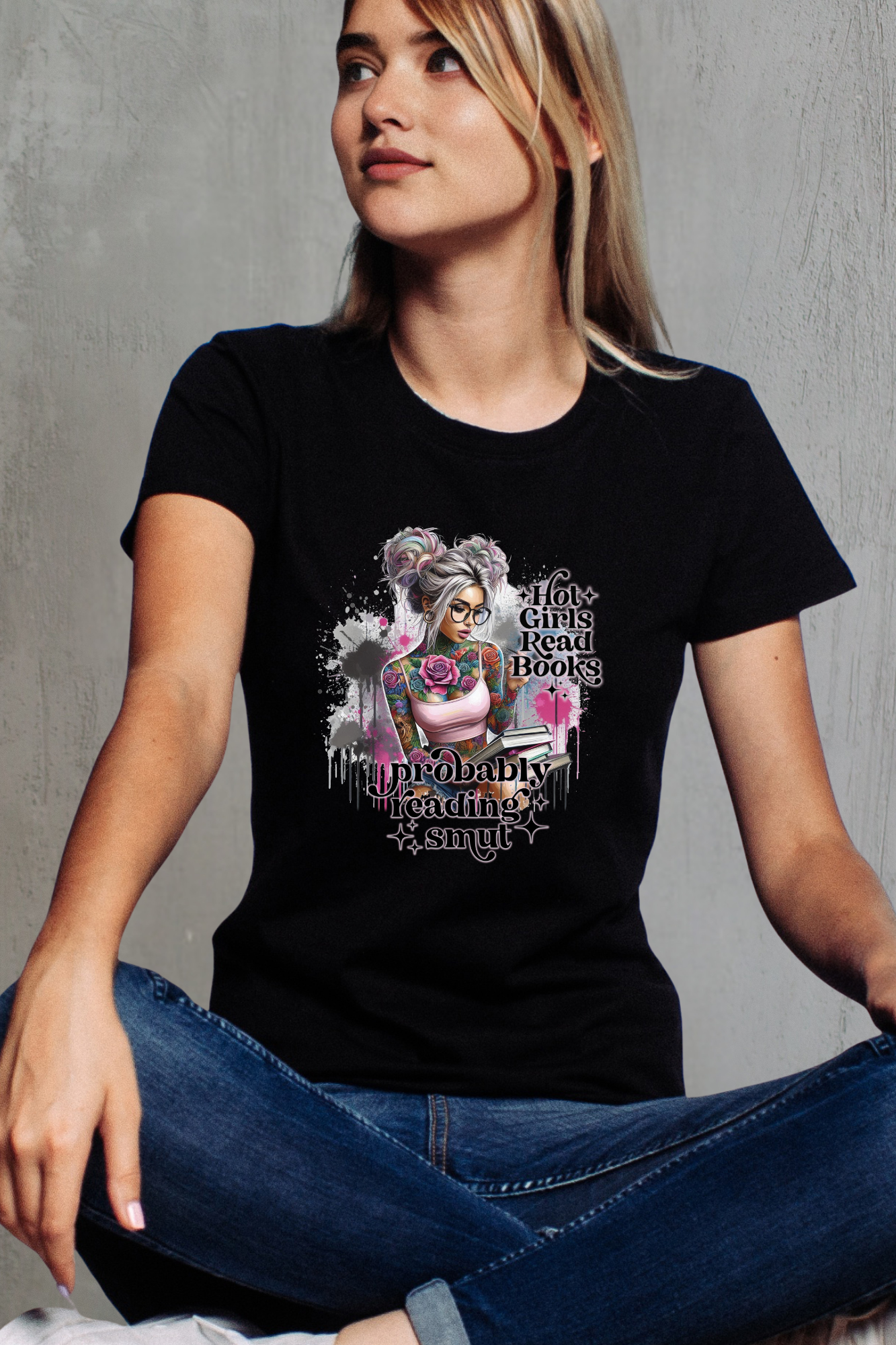 Woman wearing a black T-shirt with a colorful graphic of a tattooed girl holding books, sitting casually against a gray wall.