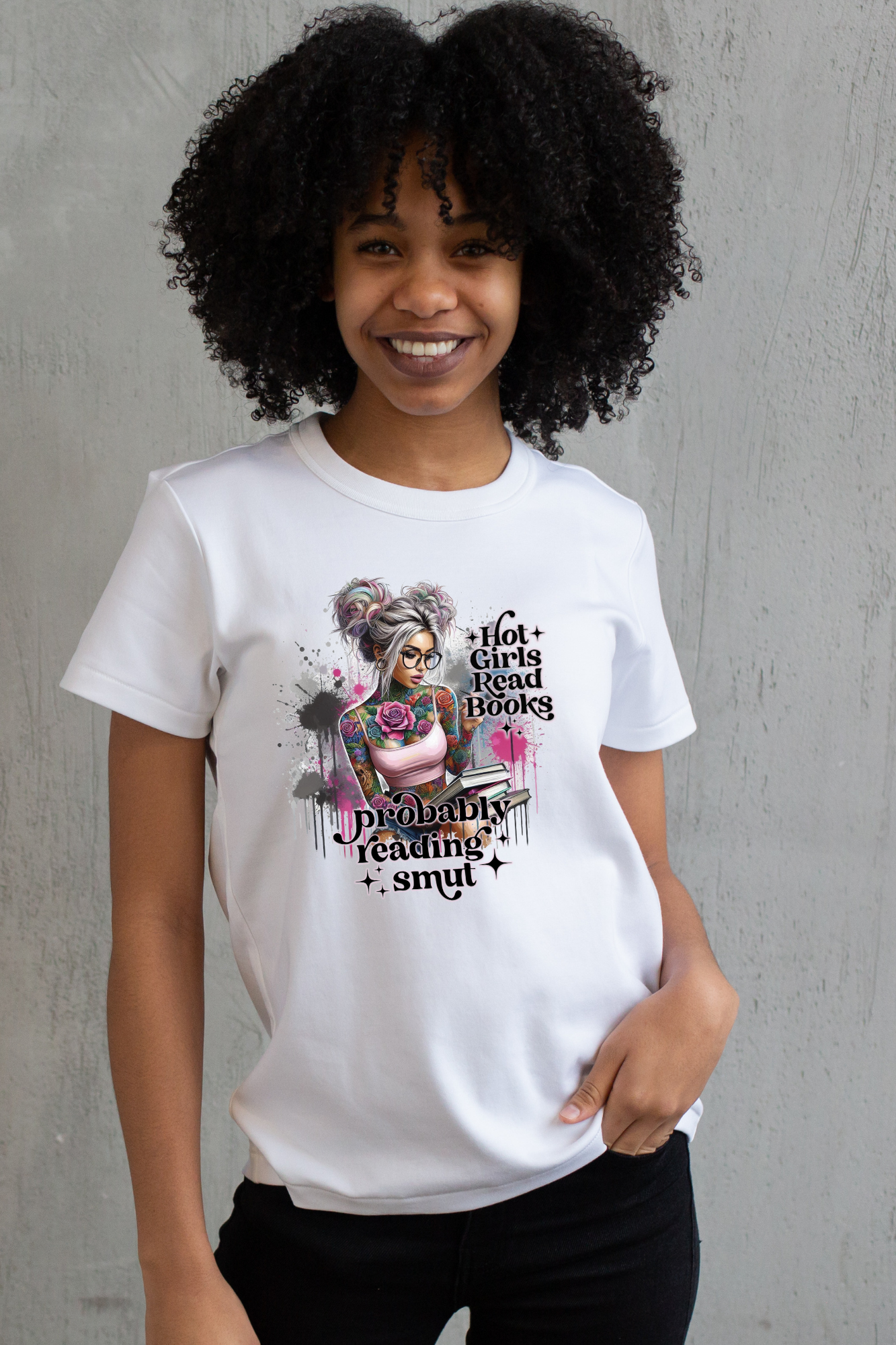 Woman wearing a white T-shirt with a colorful graphic of a tattooed girl holding books, sitting casually against a gray wall.
